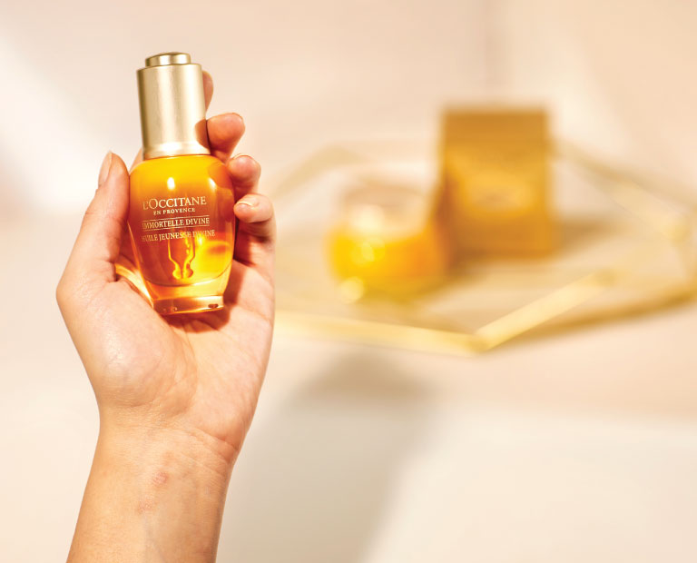A hand holding the Immortelle Divine Serum with The immortelle cream in the background