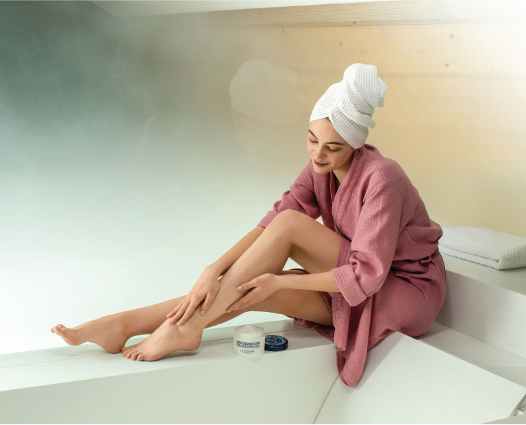 A woman dressed in a robe applying l'Occitane's Shea Butter Ultra Rich Body Cream on her legs